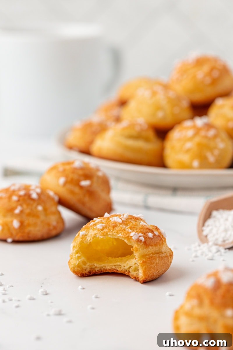A broken-open chouquette showing the light, airy center, surrounded by more chouquettes on a white surface and on a white plate in the background, emphasizing its perfect internal structure and delicate shell.