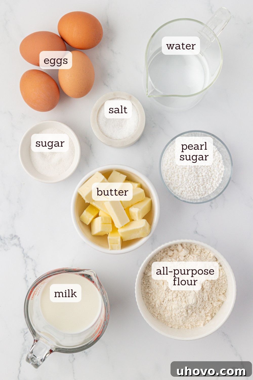 Overhead view of all the key ingredients for chouquettes laid out on a clean white surface: flour, eggs, butter, milk, water, sugar, salt, and a jar of pearl sugar, showcasing their freshness and quality.