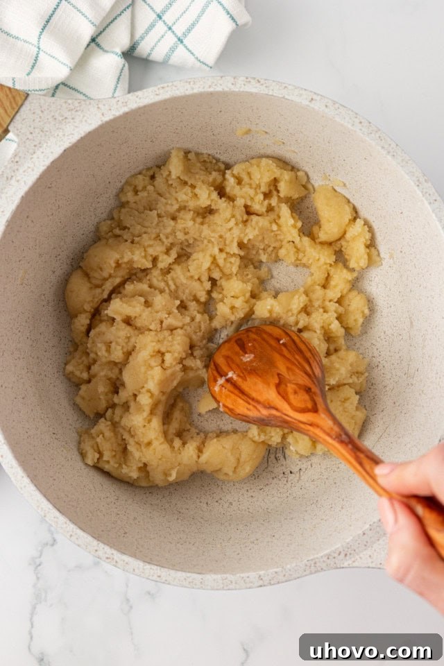 overhead view of choux pastry being vigorously stirred with a wooden spoon in a saucepan over heat, forming a cohesive ball that pulls away from the sides