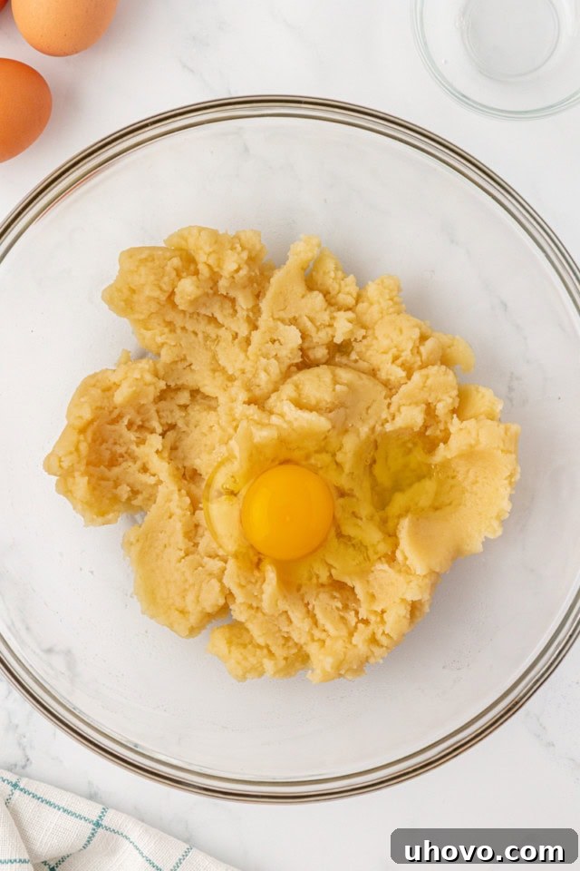 overhead view of a single egg cracked into the warm choux pastry dough in a mixing bowl, ready to be beaten in