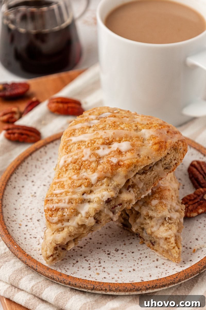 Two golden-brown maple pecan scones, beautifully drizzled with maple glaze, rest on a white plate beside a steaming mug of coffee and a small pitcher of pure maple syrup in the background, evoking a cozy fall morning.