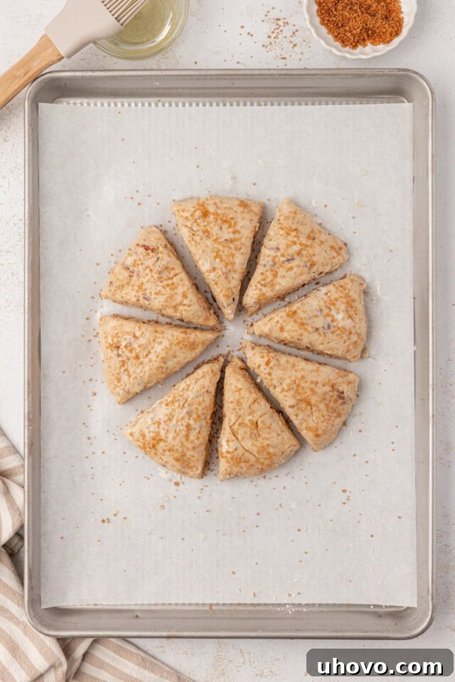 Freshly cut maple scone wedges arranged on a parchment-lined baking sheet, brushed with egg white and sprinkled with coarse sugar, ready for the oven.