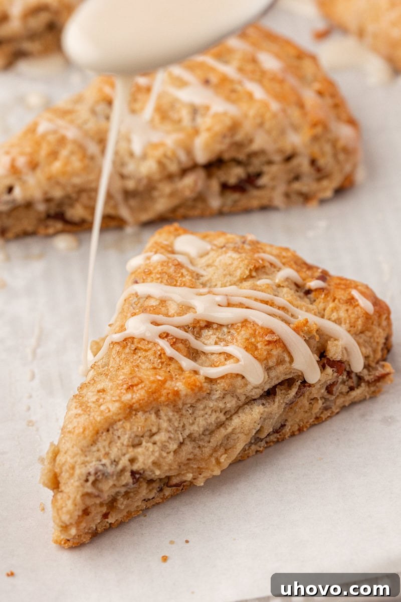 A close-up shot of a single maple scone being expertly drizzled with a thick, glossy maple glaze, highlighting its golden-brown crust and moist texture.