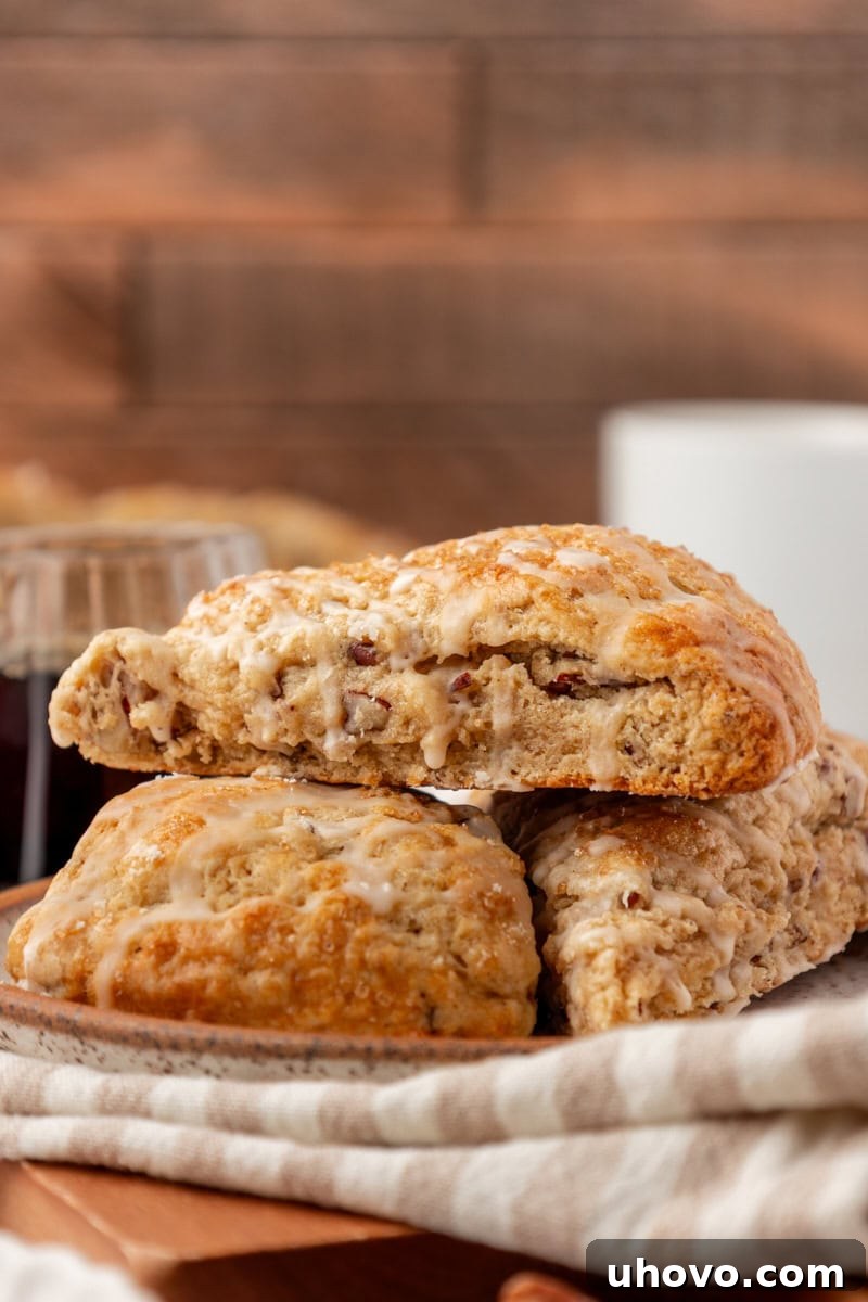 Three maple pecan scones on a white plate, positioned next to a mug of steaming coffee and a small pitcher of maple syrup, creating an inviting breakfast scene.
