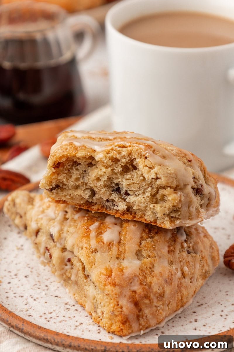 A close-up shot of a maple pecan scone with a bite taken out, revealing its tender, flaky interior, propped on top of another scone on a white plate.