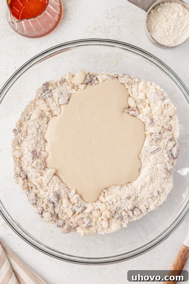 Wet ingredients (Greek yogurt, egg yolk, maple syrup) are being poured into the bowl of dry ingredients (flour, butter crumbs, pecans) for maple scones.