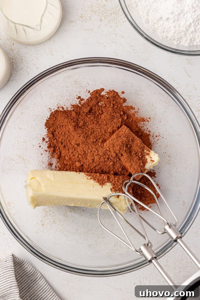 An overhead view showing softened butter and unsweetened cocoa powder combined in a clear glass mixing bowl, ready to be beaten together for the chocolate frosting.