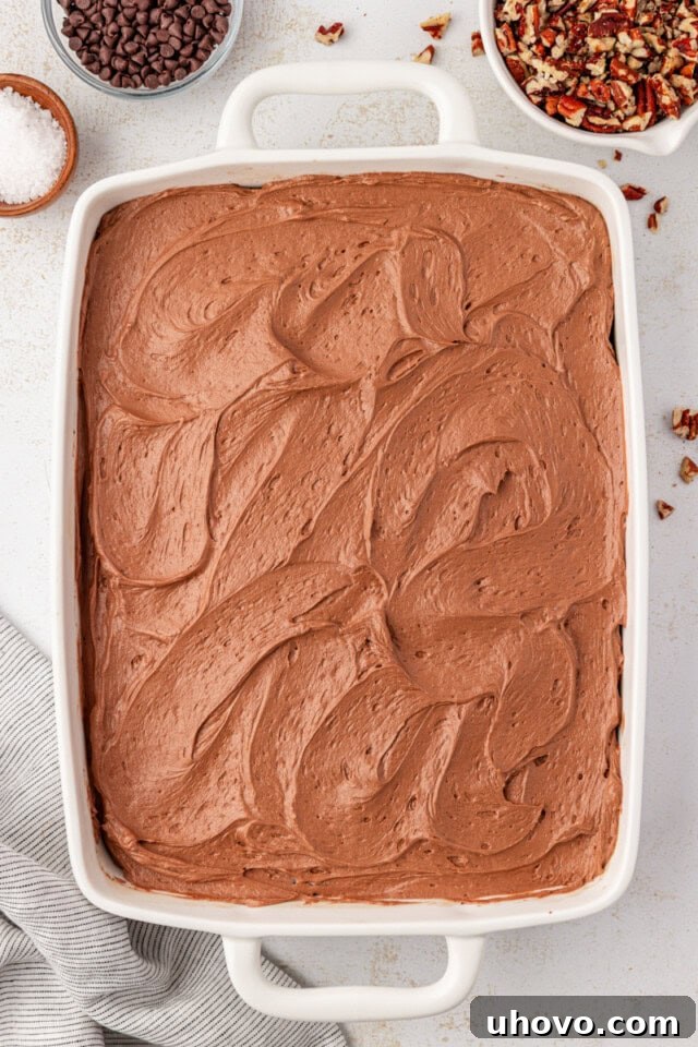 An overhead view of rich chocolate frosting being smoothly spread over the completely cooled chocolate caramel cake in a white rectangular baking pan.