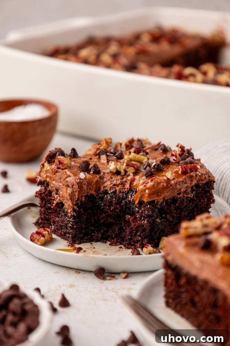 A close-up view of a single slice of turtle cake on a white plate, with a small bite taken out, revealing the moist chocolate cake, gooey caramel, and speckled pecans and chocolate chips, with more cake blurred in the background.
