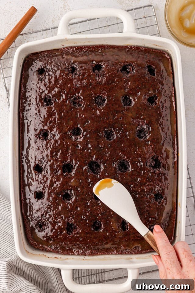 An overhead view capturing the moment warm caramel sauce is being spread over the surface of the chocolate cake, which has been strategically poked with holes to absorb the filling.