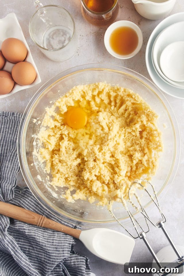 Overhead view of a single egg being added to the sandy cake batter mixture in a glass bowl.