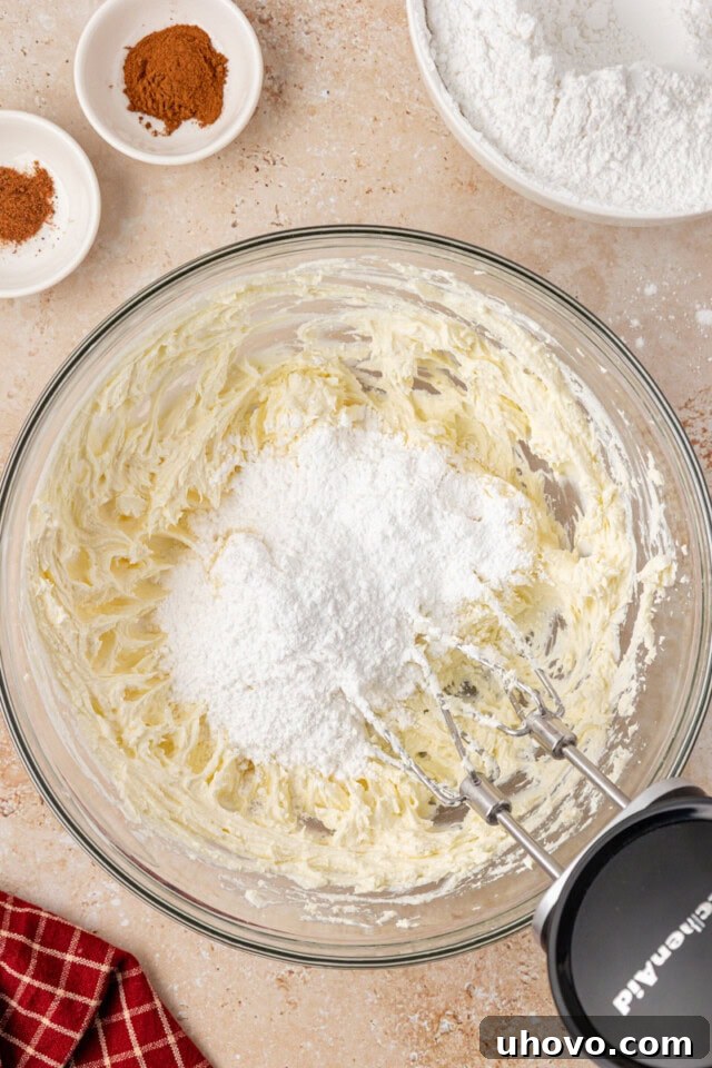 Overhead view of confectioners' sugar being slowly added to a bowl of creamed butter and cream cheese, preparing the base for spiced cream cheese frosting.