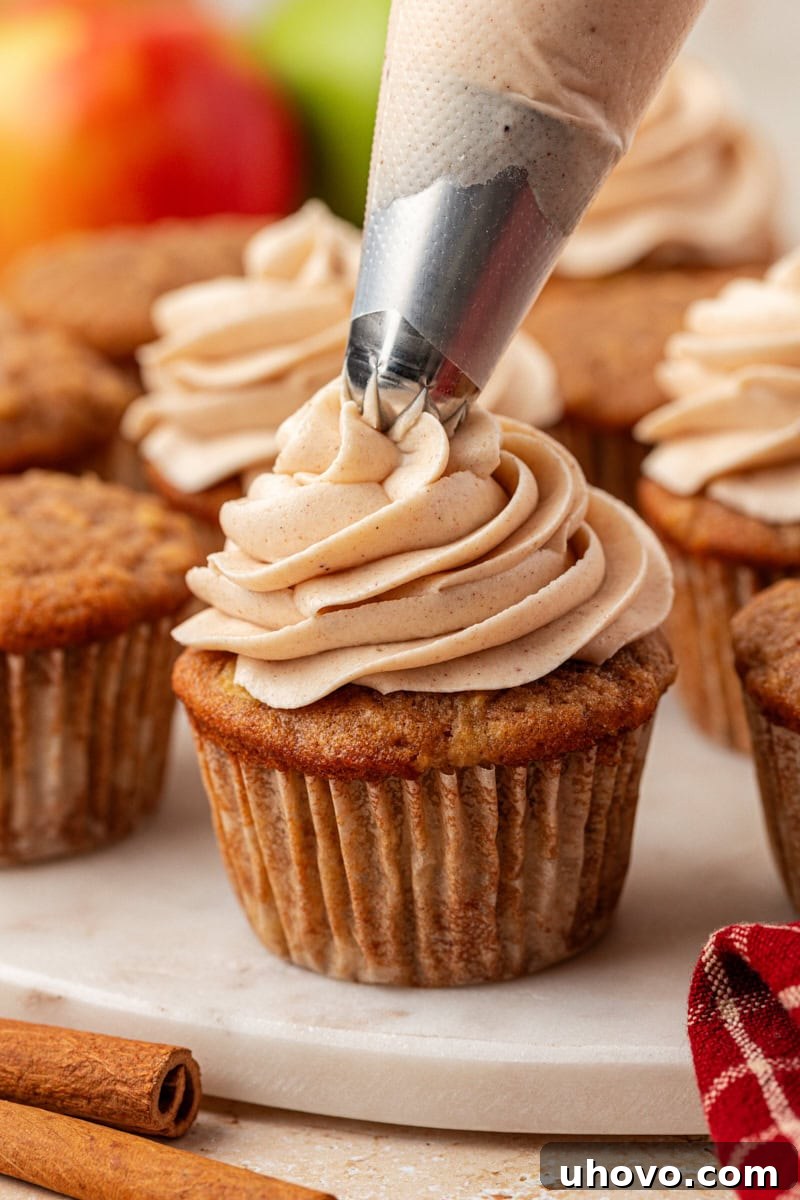 A close-up shot of creamy spiced cream cheese frosting being expertly piped onto a freshly baked apple cupcake, with more tempting cupcakes visible in the soft-focus background.