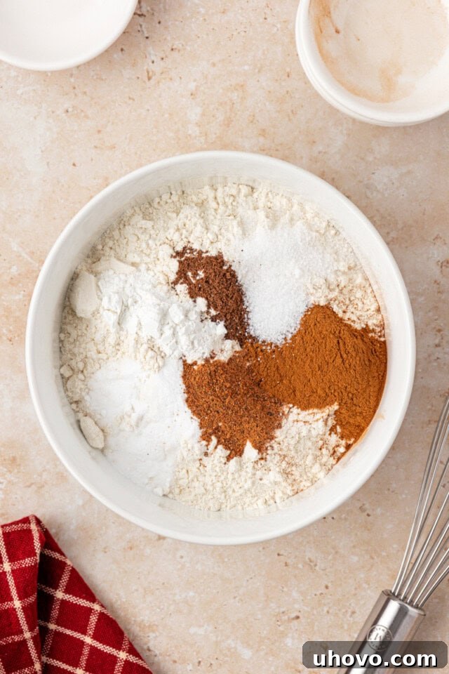 Overhead view of flour, baking powder, baking soda, salt, and various warm spices meticulously combined in a white mixing bowl, ready for the next step in making apple cupcakes.