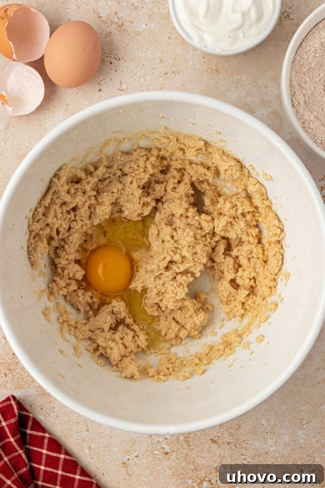 Close-up, overhead view of a single egg being cracked and added to a bowl of creamed butter and sugar, being prepared for apple cupcakes.