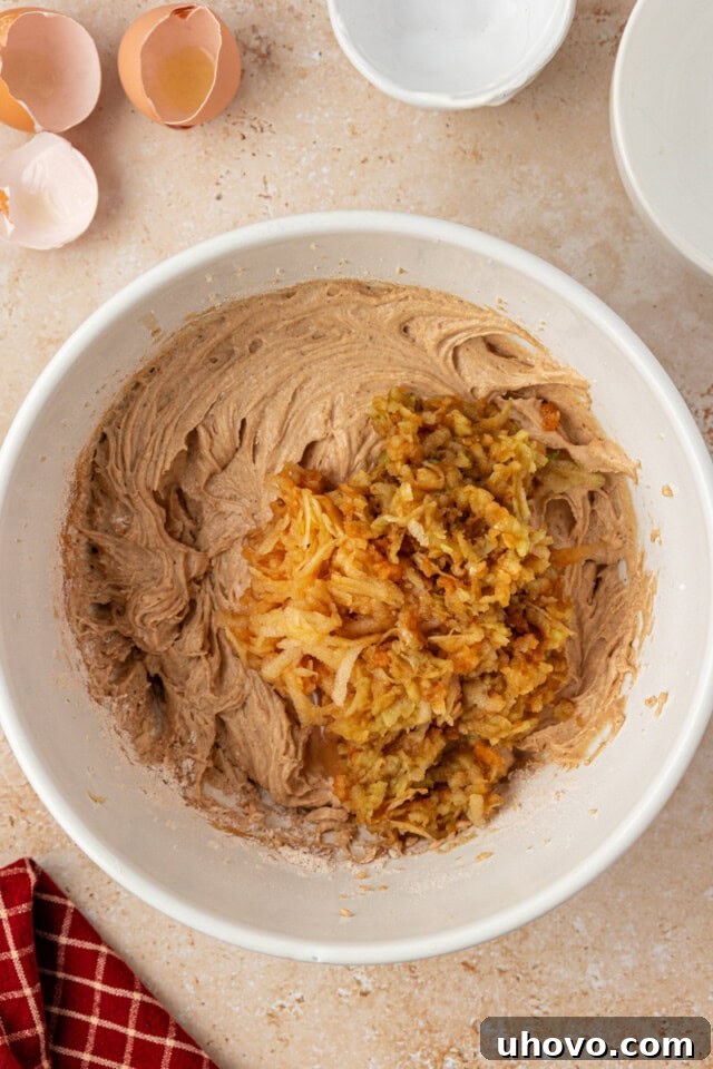 Overhead close-up of freshly grated apples being gently folded into the thick, spiced cupcake batter using a spatula, ensuring even distribution.