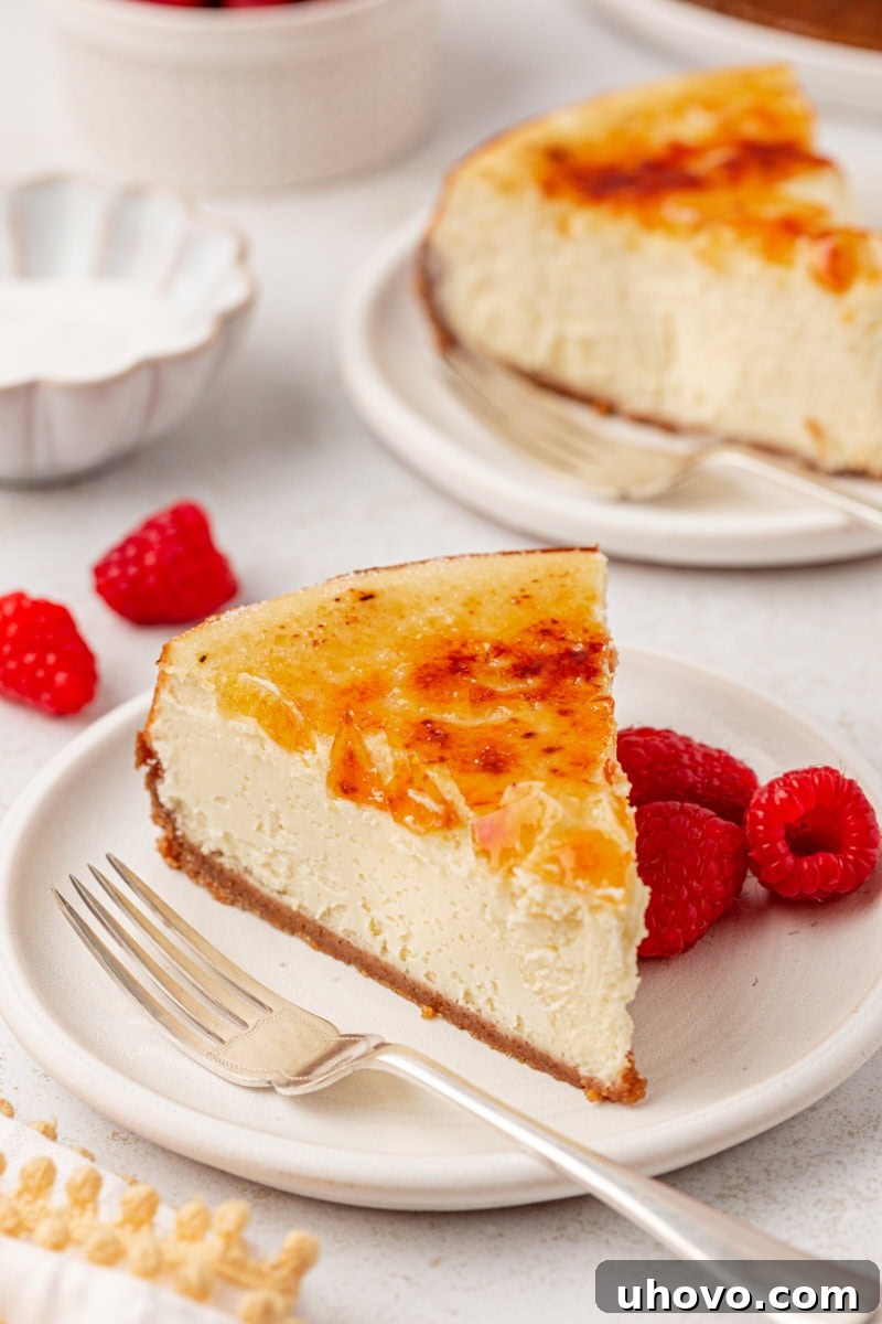 a slice of crème brûlée cheesecake on a white plate with another slice and fresh raspberries in the background