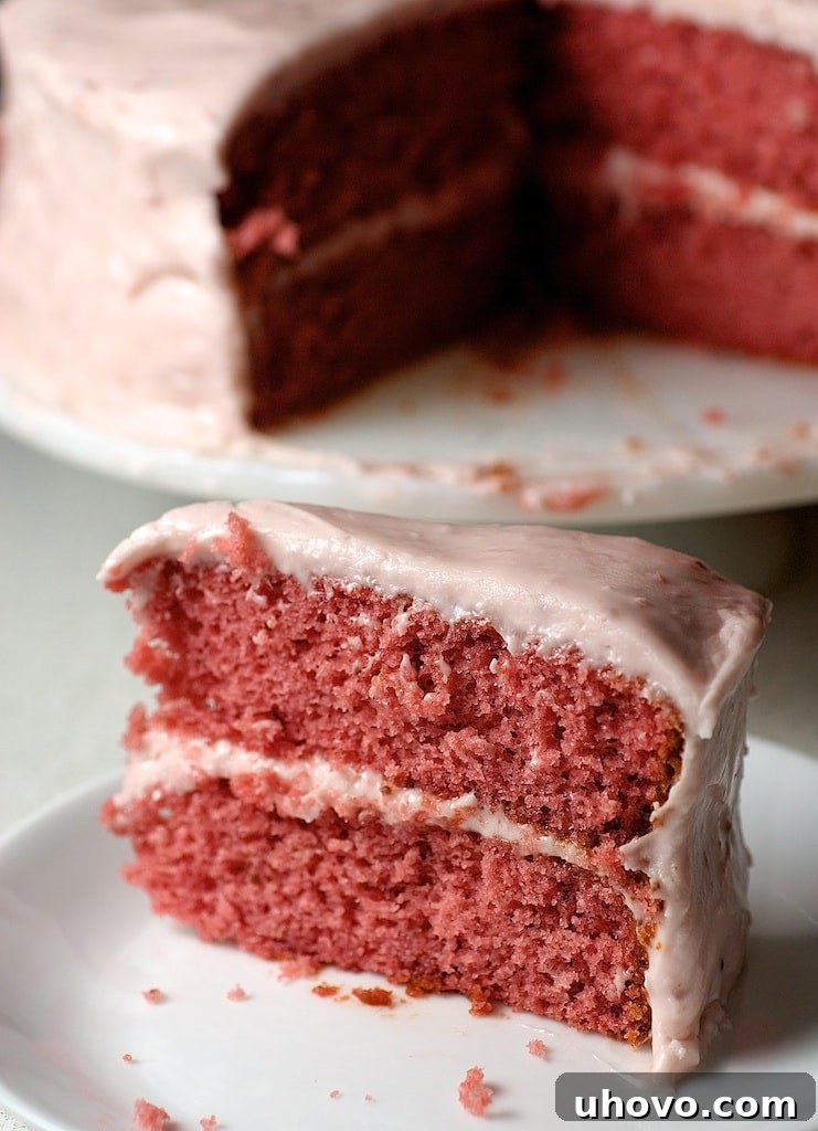A beautifully presented slice of strawberry cake on a white plate, with the remaining layered cake visible in the soft-focus background, highlighting its fluffy texture and vibrant pink frosting.