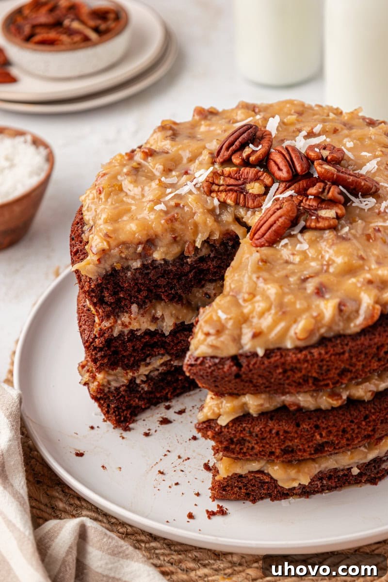 German chocolate cake on a white cake plate with a slice missing, showcasing the rich chocolate layers and abundant, creamy coconut-pecan frosting.