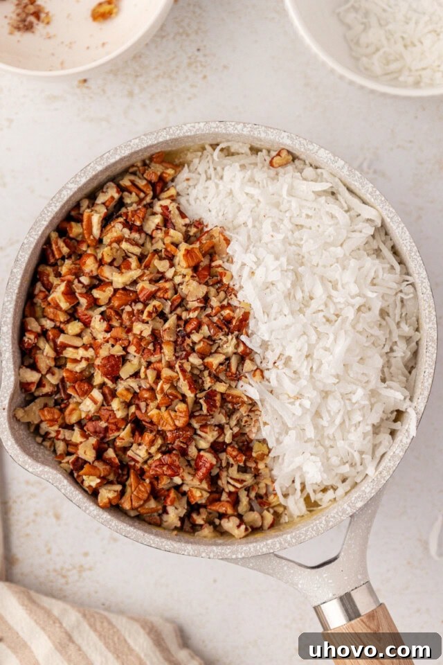Overhead view of sweetened shredded coconut, chopped pecans, and vanilla extract being stirred into the cooked, warm frosting mixture in a saucepan.