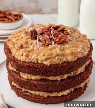 German chocolate cake on a white cake plate, with a slice removed, displaying the layers and rich coconut-pecan frosting.