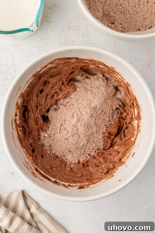 Overhead view of buttermilk being added to the chocolate cake batter in a mixing bowl, alternating with dry ingredients for a balanced consistency.