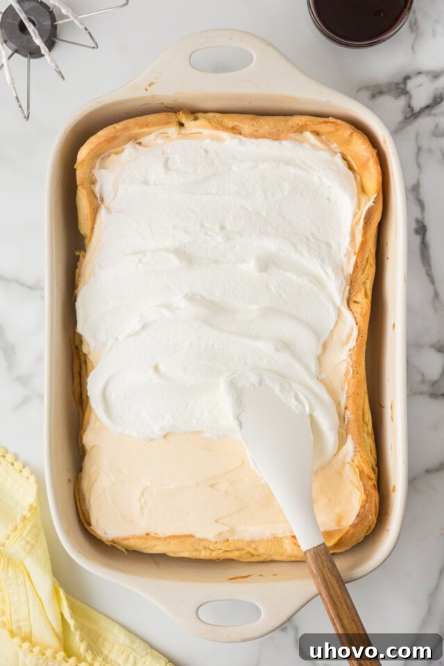 Overhead view of additional freshly whipped cream being spread generously over the vanilla filling layer of the Cream Puff Cake in a white rectangular pan.