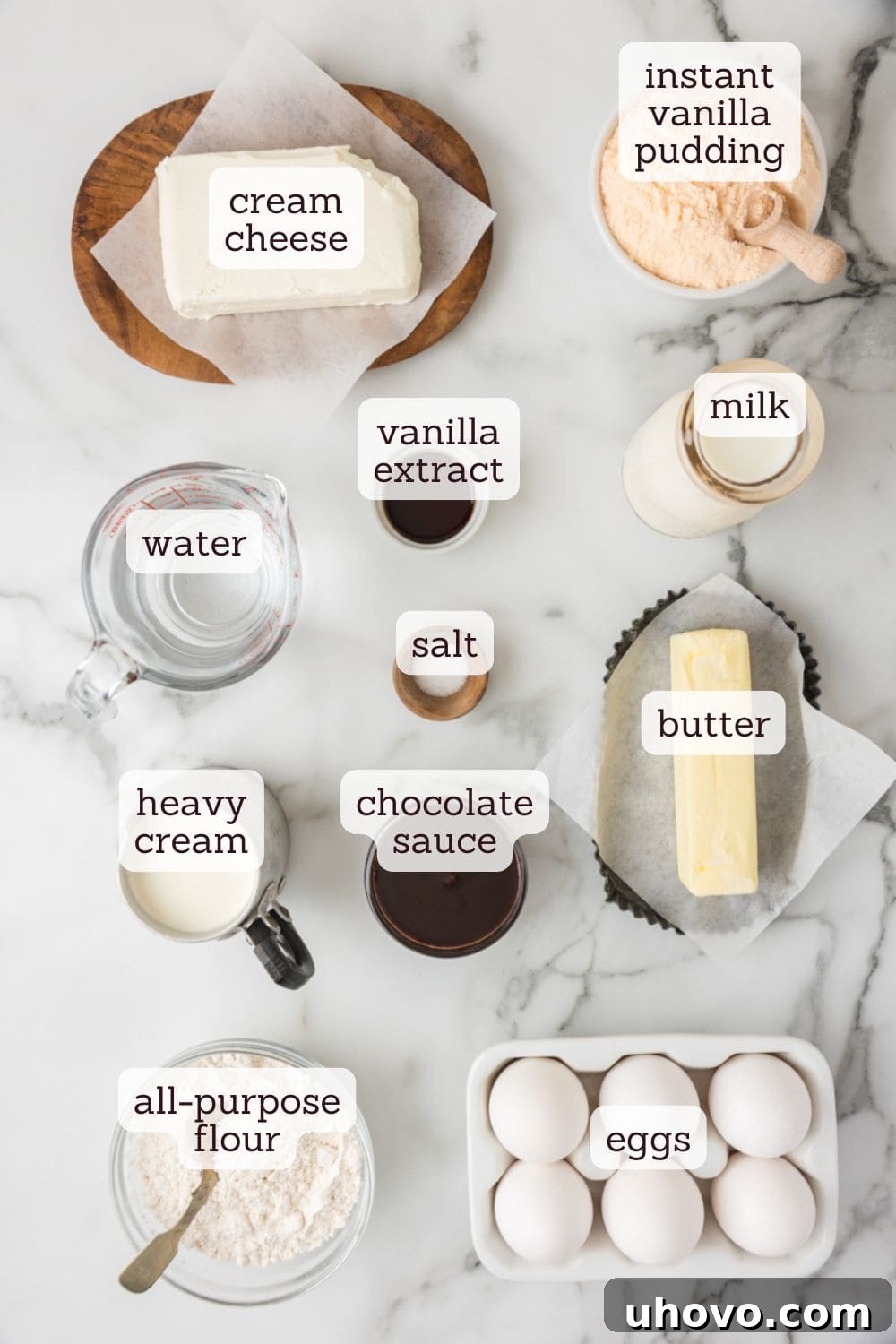 Overhead view of various ingredients neatly arranged on a counter, including butter, eggs, flour, milk, cream cheese, and pudding mix, prepared for making a Cream Puff Cake.