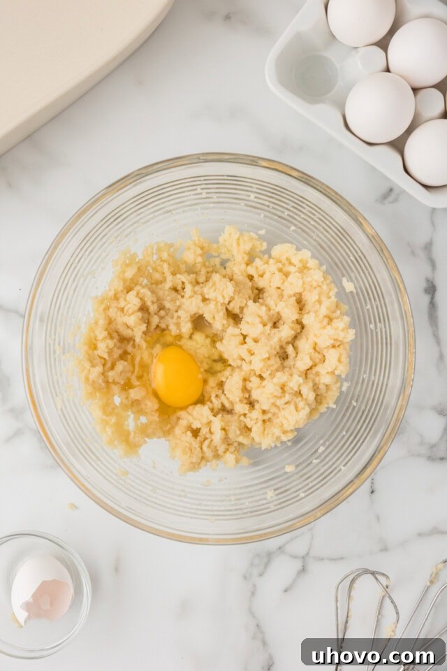 Close-up overhead shot of an egg being added to the warm choux pastry mixture in a bowl, ready for beating to incorporate.