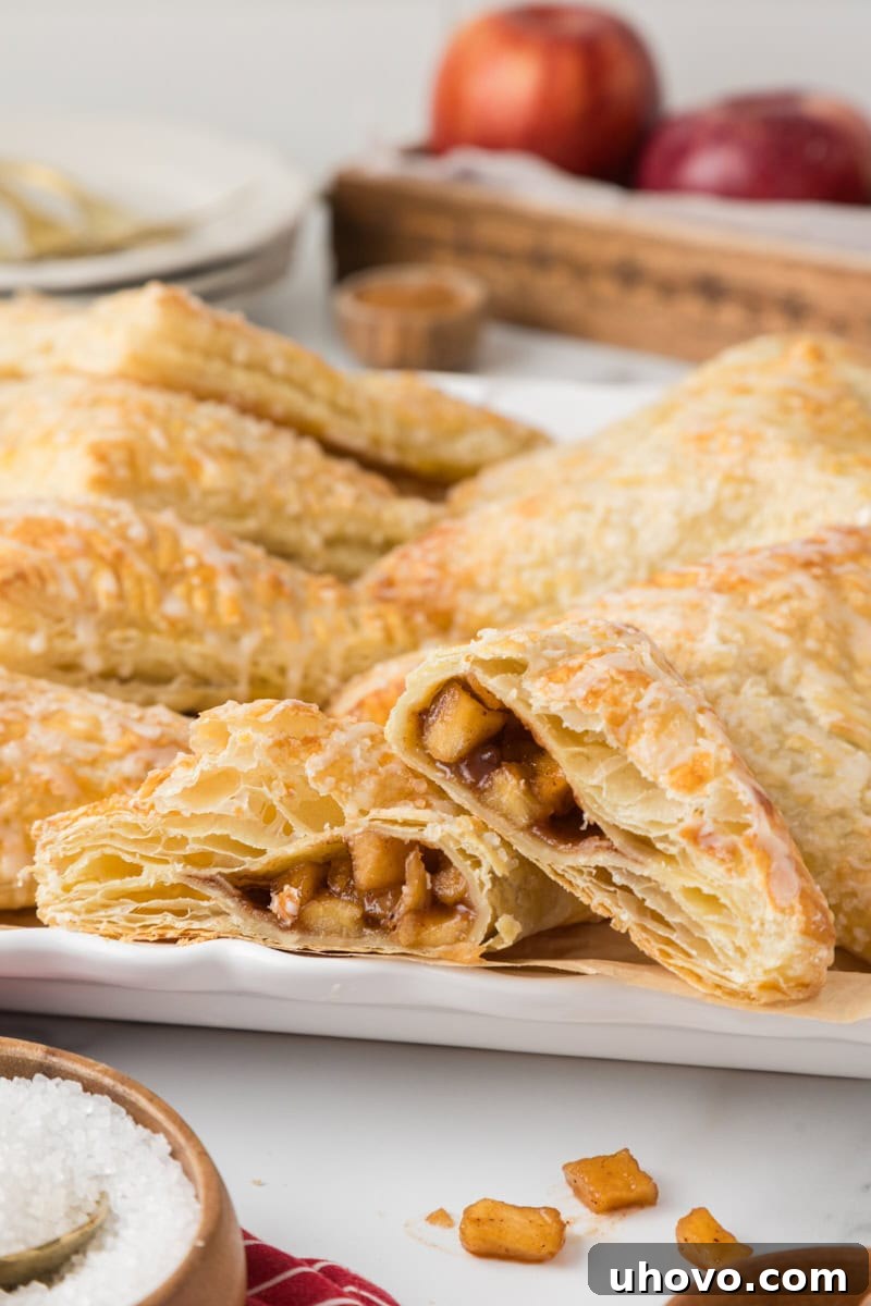Several golden brown apple turnovers on a white plate, with one turnover cut in half to show the sweet apple filling and flaky layers.