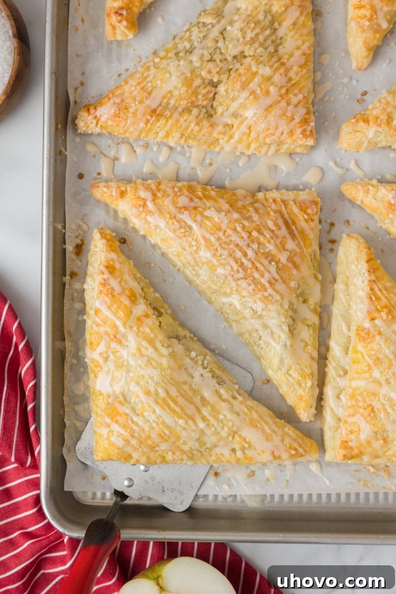 Overhead view of freshly glazed apple turnovers resting on a parchment-lined baking sheet, their golden crusts glistening.