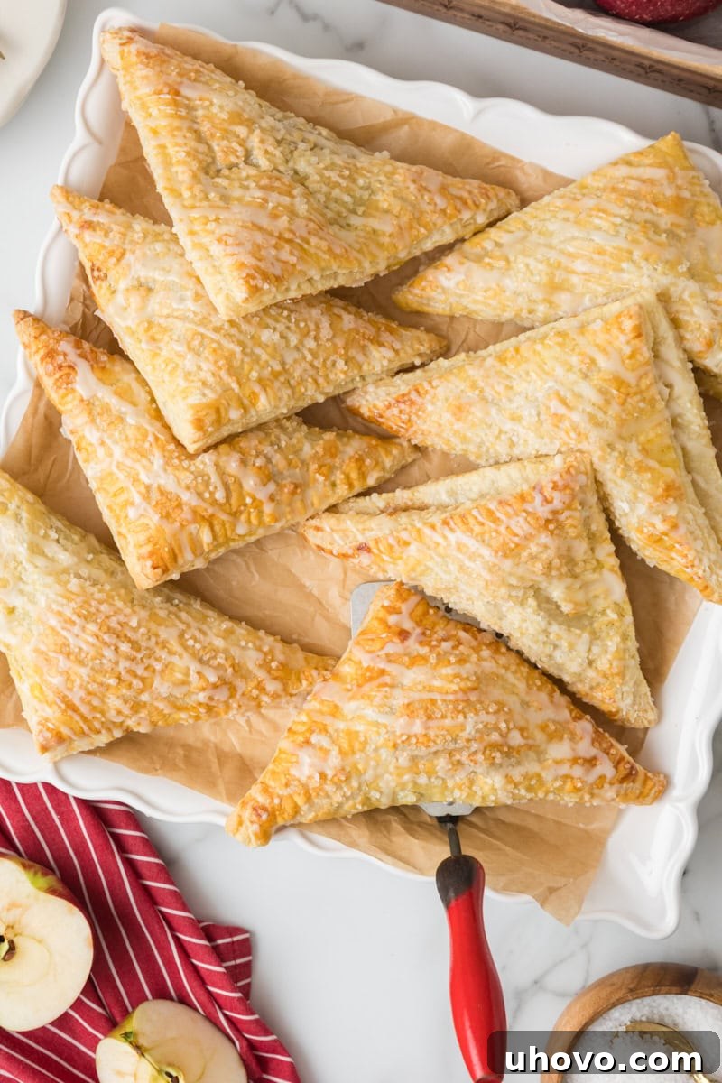 Overhead view of two perfectly baked and glazed apple turnovers served on a square white plate, with more in the background.