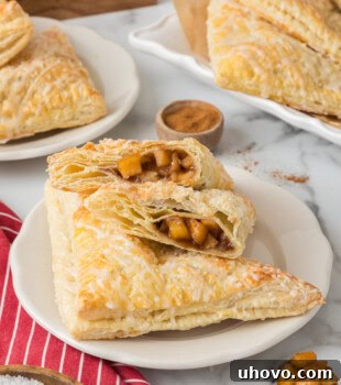 Two halves of an apple turnover along with another whole turnover on a white plate with more pastries in the background, showing flaky pastry and apple filling.