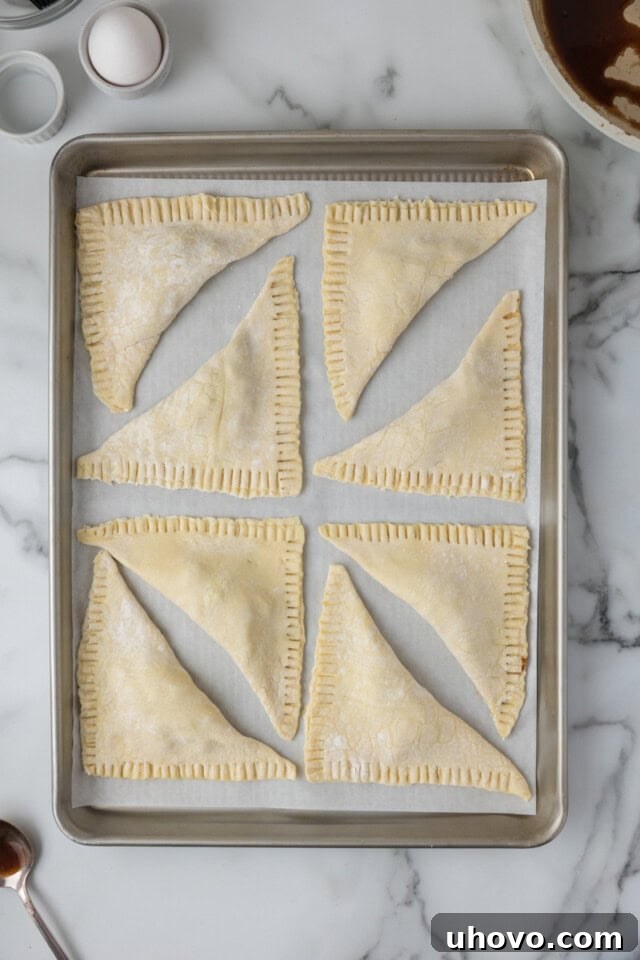 Overhead view of assembled apple turnovers, folded into triangles, sealed, and arranged on a parchment-lined baking sheet, ready for chilling.