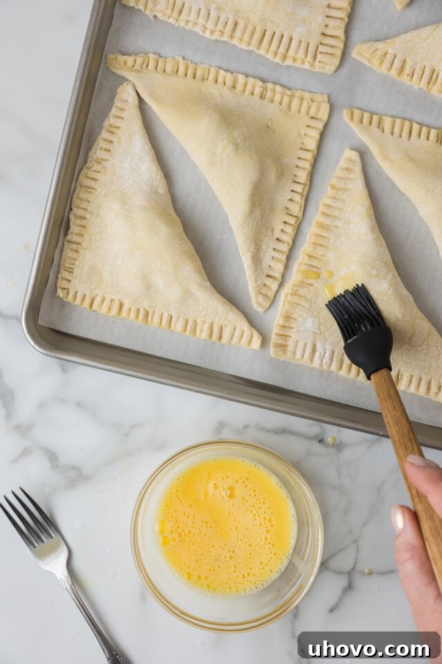 Overhead view of an egg wash being delicately brushed over unbaked apple turnovers on a sheet pan, ready for the oven.