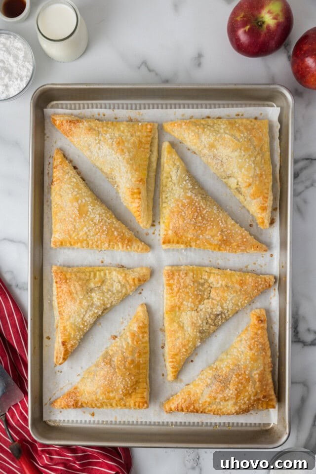 Overhead view of freshly baked, golden brown apple turnovers cooling on a sheet pan, visibly puffed and flaky.