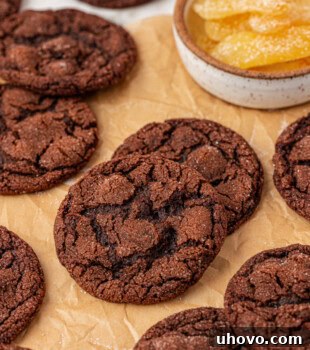 Several chocolate ginger cookies scattered on parchment paper with a bowl of candied ginger in the background, creating an enticing scene.