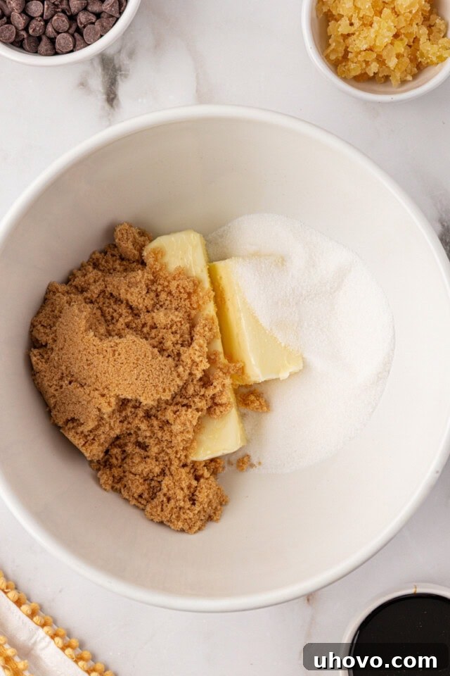 An overhead view of softened butter, granulated sugar, and light brown sugar being creamed together in a white mixing bowl, forming a pale, fluffy mixture.