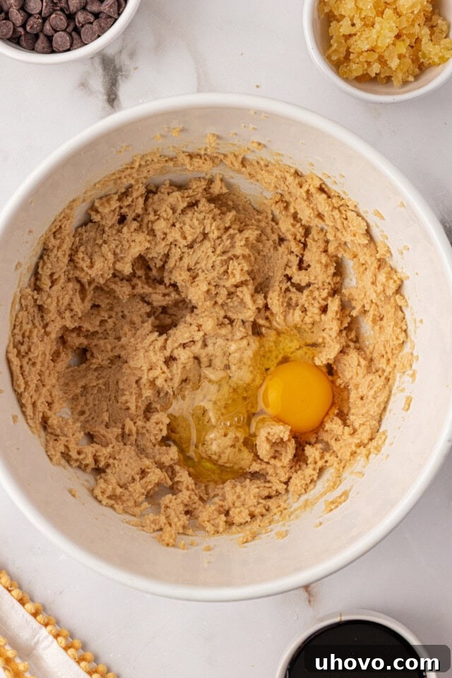 An overhead view of an egg freshly added to the creamy mixture of butter and sugars in a mixing bowl, ready to be incorporated.