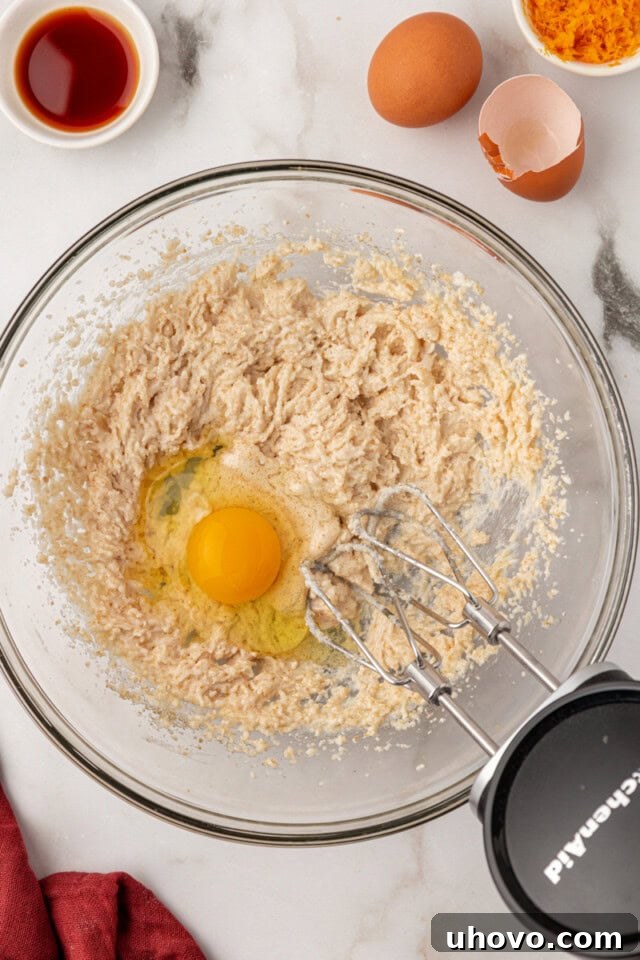 Overhead view of a single egg being added to the creamed butter, oil, and sugar mixture in a glass bowl, followed by vanilla and orange zest.