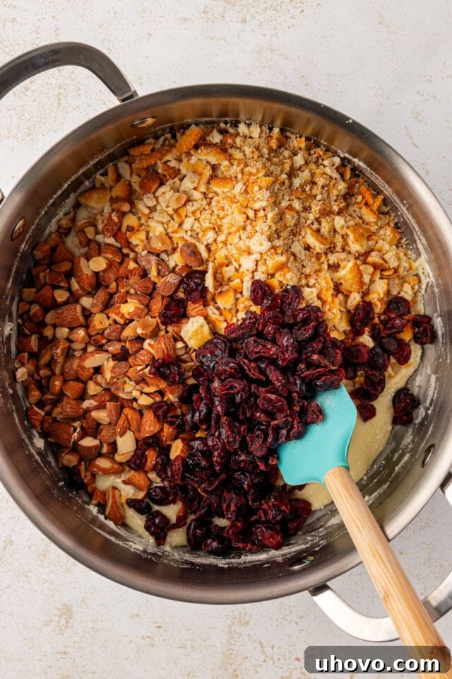 Overhead view of coarsely crushed crackers, dried cranberries, and chopped nuts being added to the warm Snowflake Crisp base mixture in a large saucepan.