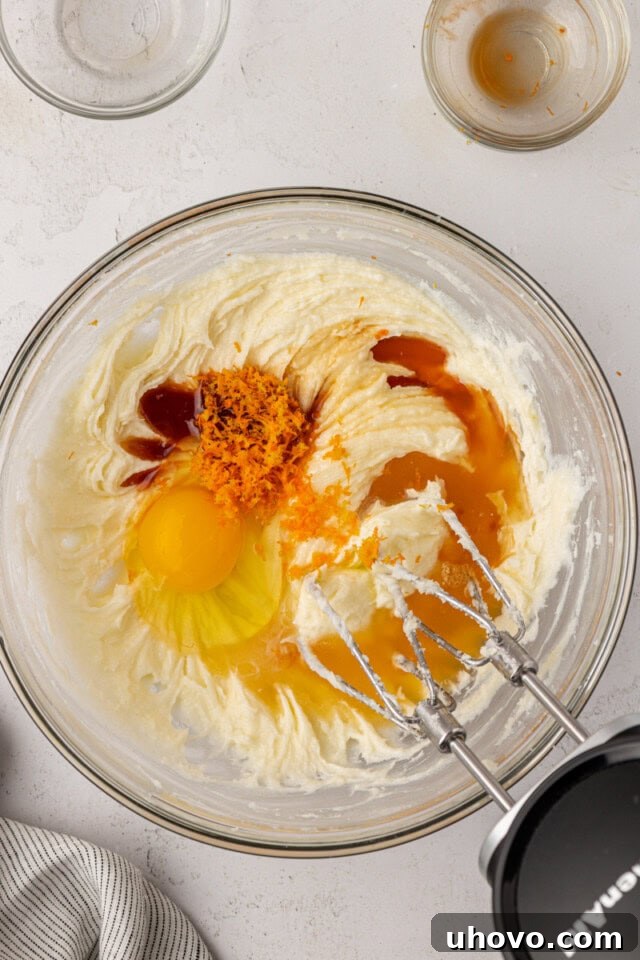 overhead view of egg, orange zest, orange juice, vanilla, and almond extract added to creamed butter, cream cheese, and sugar in a mixing bowl