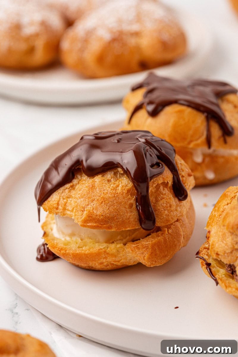 A perfectly golden profiterole, split open and filled with creamy vanilla ice cream, then generously topped with glistening warm chocolate sauce, resting on a pristine white plate.