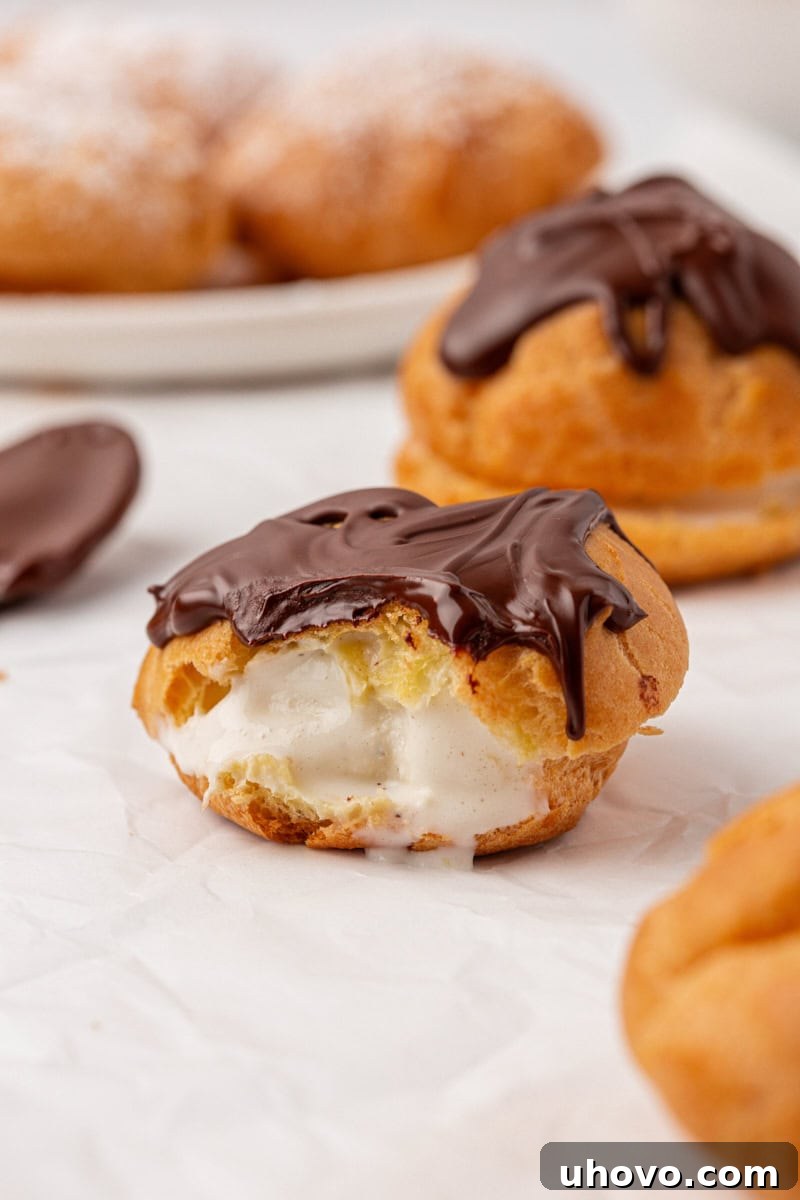 A close-up of an ice cream-filled profiterole with a bite taken out, revealing the creamy interior and hollow choux pastry, resting on a white plate.