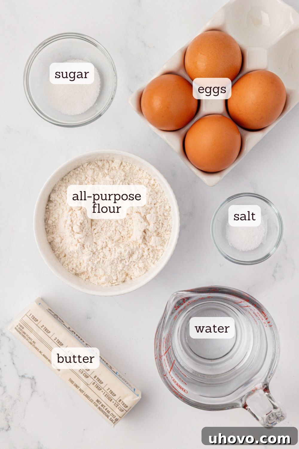 An overhead view showcasing the essential ingredients for classic profiteroles neatly arranged: butter, water, sugar, salt, flour, and eggs, along with a whisk and measuring cups.