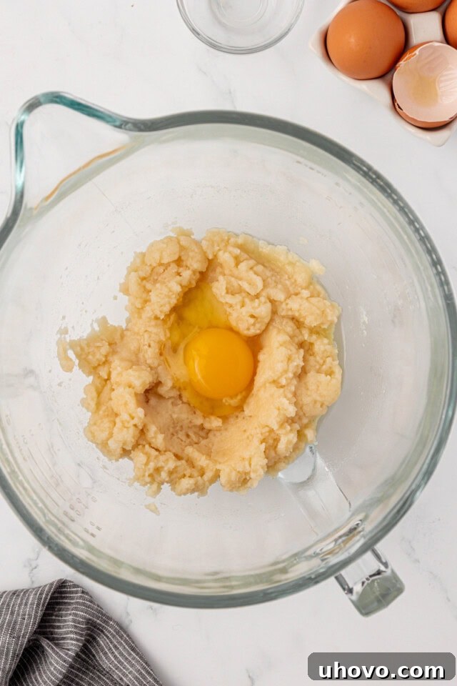 A hand adding a beaten egg to the slightly cooled pâte à choux dough in a mixing bowl, prior to full incorporation.