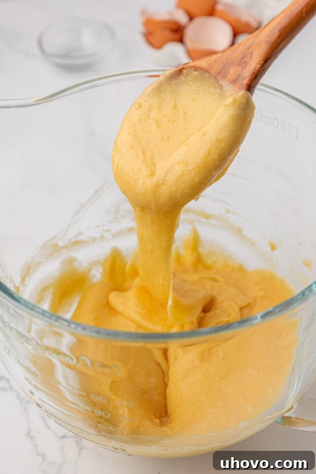 A wooden spoon lifting a thick, glossy ribbon of pâte à choux dough, demonstrating the perfect consistency.