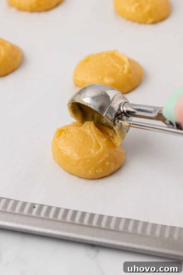 Pâte à choux dough being neatly portioned onto a parchment-lined baking sheet using a cookie scoop, ready for baking.