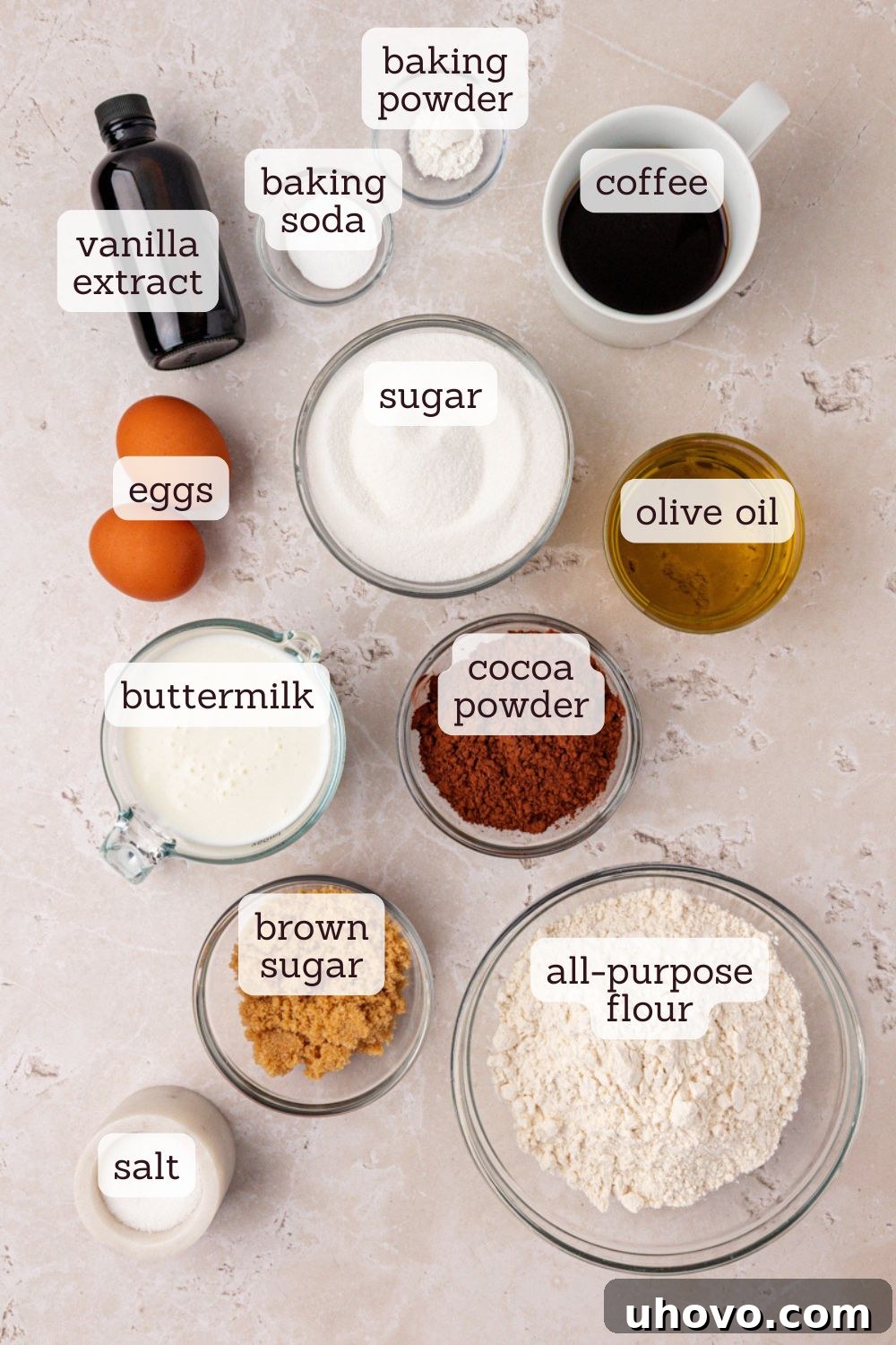 An overhead view of all the key ingredients for chocolate olive oil cake laid out on a surface, including olive oil, cocoa powder, flour, sugar, eggs, and buttermilk.