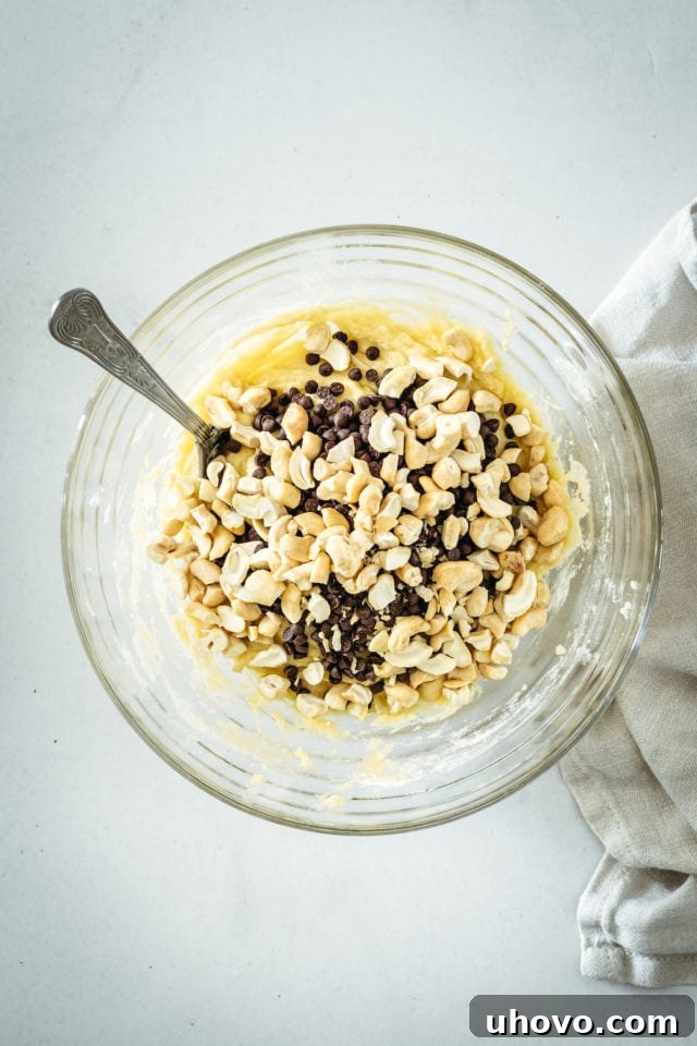 An overhead view of chocolate chips and chopped cashews being gently stirred into the bread batter in a glass mixing bowl, ensuring even distribution.